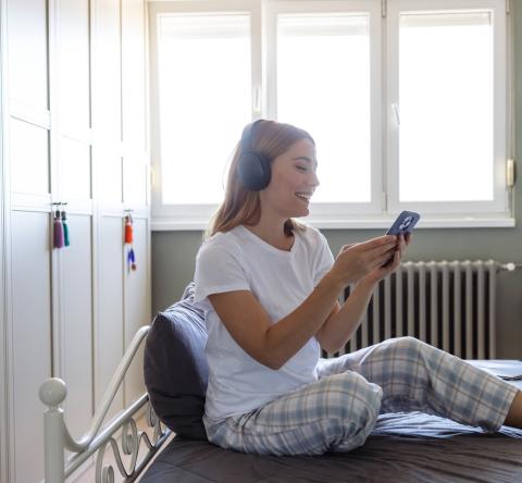 Une jeune femme blonde en pyjama regarde son téléphone, assise sur son lit avec un casque audio sur les oreilles