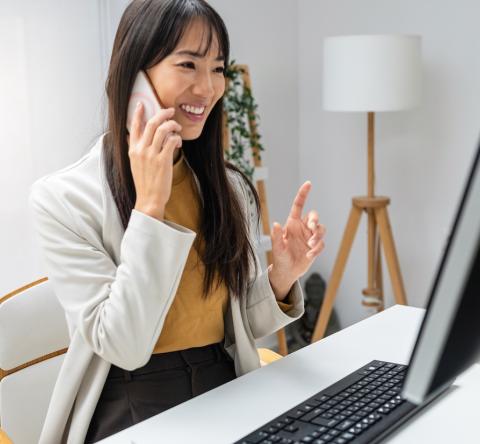 Une femme aux cheveux longs est en pleine conversation téléphonique, assise devant son ordinateur 
