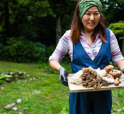 Une femme marche dans son jardin, en portant une planche en bois avec les champignons qu'elle vient de ramasser