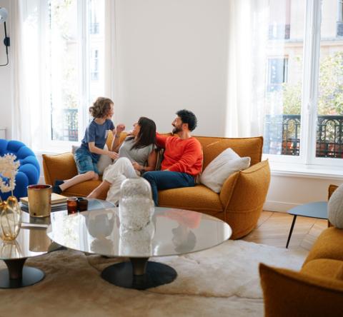 Un couple et un enfant rigolent assis sur un canapé jaune moutarde dans un bel appartement parisien. 