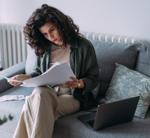 Femme travaillant sur son canapé gris, lisant ses documents, avec son ordinateur posé non loin. 